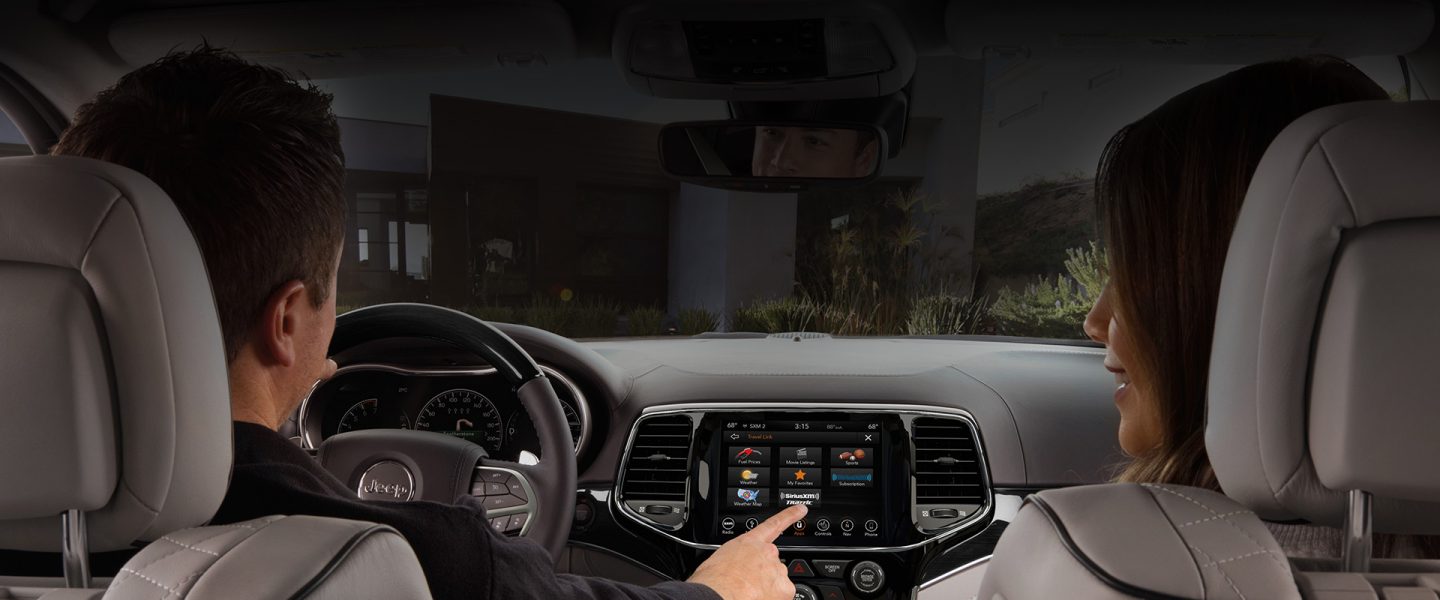 A couple in the front seat of the 2020 Jeep Grand Cherokee as the driver makes a selection on the Uconnect touchscreen.